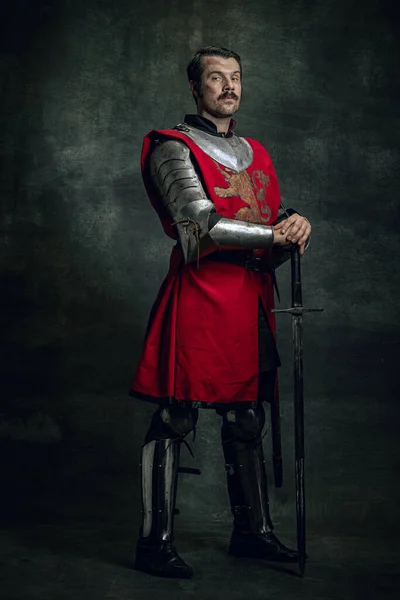 Retrato de seriuos brutais homem, guerreiro medieval ou cavaleiro com rosto sujo ferido segurando espada isolada sobre fundo escuro. Comparação de eras — Fotografia de Stock