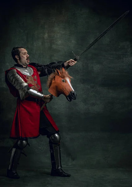 Retrato de guerreiro medieval ou cavaleiro usando capacete e armadura cavalo de brinquedo, segurando grande espada isolada sobre fundo escuro. Comparação de eras, história — Fotografia de Stock