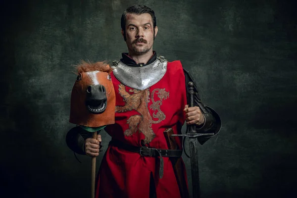 Porträt eines mittelalterlichen Kriegers oder Ritters mit Helm und Rüstung, der Spielzeugpferd reitet und ein großes Schwert isoliert vor dunklem Hintergrund hält. Vergleich von Epochen, Geschichte — Stockfoto