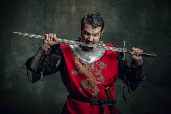 Komický portrét legračního středověkého válečníka nebo rytíře se špinavou zraněnou tváří držícího meč izolovaný na tmavém pozadí. Porovnání období — Stock fotografie