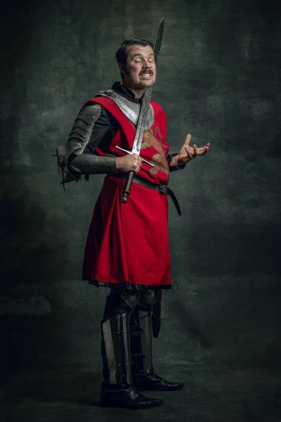 Комічний портрет смішного середньовічного воїна або лицаря з брудним пораненим обличчям, що тримає меч ізольовано на темному тлі. Порівняння епох — стокове фото