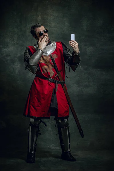 Jovem sorridente em imagem de guerreiro medieval ou cavaleiro com rosto sujo ferido usando telefone isolado sobre fundo escuro. Comparação de eras — Fotografia de Stock