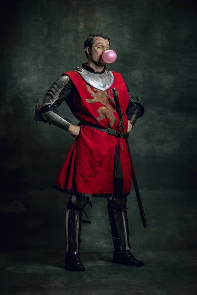 Komisches Porträt eines lustigen mittelalterlichen Kriegers oder Ritters mit schmutzigem, verwundetem Gesicht, das Schwert isoliert vor dunklem Hintergrund hält. Epochenvergleich — Stockfoto