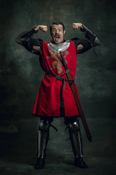 Κωμικό πορτραίτο αστείου μεσαιωνικού πολεμιστή ή ιππότη με βρώμικο πληγωμένο πρόσωπο που κρατά το σπαθί απομονωμένο σε σκοτεινό φόντο. Σύγκριση εποχών — Φωτογραφία Αρχείου