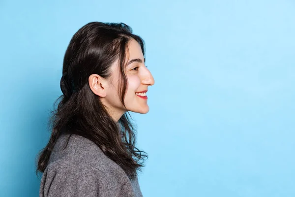 Visão de perfil da jovem menina indiana alegre vestindo pulôver cinza isolado no fundo azul. Conceito de emoções, expressão facial, juventude, aspiração, vendas — Fotografia de Stock