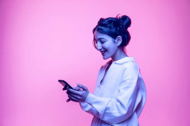 Beyaz bluzlu güzel bir kızın pembe arka planda neon ışıkta izole edilmiş akıllı telefon kullanarak çekimleri. Duyguların kavramı