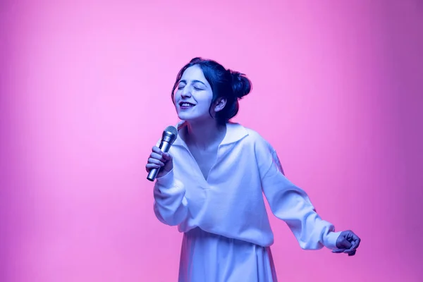Retrato de jovem bela menina sorridente em blusa branca cantando no microfone isolado no fundo rosa em néon. Conceito de emoções — Fotografia de Stock