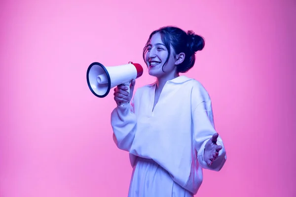 Joven hermosa chica feliz en blusa blanca gritando a megáfono aislado sobre fondo rosa en luz de neón. Concepto de emociones —  Fotos de Stock