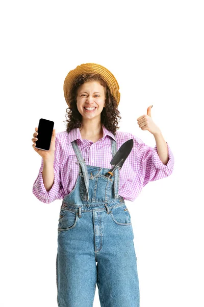 Studio girato di giovane ragazza felice, giardiniere donna in uniforme da lavoro e cappello utilizzando telefono isolato su sfondo bianco. Concetto di lavoro, emozioni, agronomia, eco — Foto Stock