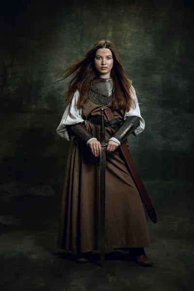Full längd porträtt av seriuos vacker kvinna i bild av medeltida krigare eller riddare med smutsiga sårade ansikte med stort svärd isolerad över mörk vintage bakgrund. — Stockfoto