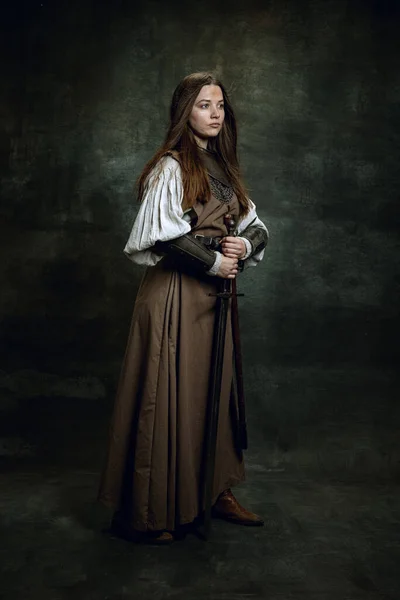 Portret pełnej długości seriuos piękna kobieta w obrazie średniowiecznego wojownika lub rycerza z brudną ranną twarzą z dużym mieczem odizolowane nad ciemnym tle vintage. — Zdjęcie stockowe