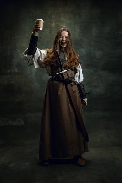 Jämförelse av epoker. Vintage porträtt av bedårande medeltida kvinna, kvinnlig krigare eller riddare med smutsigt sårat ansikte provsmakning kaffe isolerad över mörk retro bakgrund. — Stockfoto