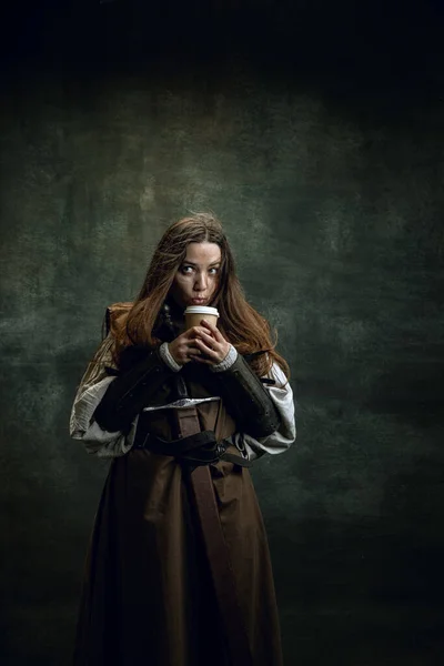 Epochenvergleich. Vintage-Porträt einer entzückenden mittelalterlichen Frau, Kriegerin oder Ritterin mit schmutzigem, verwundetem Gesicht, die vor dunklem Retro-Hintergrund isoliert Kaffee probiert. — Stockfoto