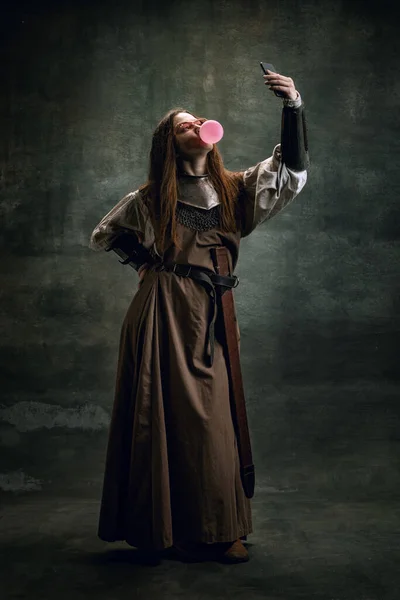 Retrato vintage de una joven hermosa en imagen de una mujer medieval guerrera usando un gadget moderno aislado sobre un fondo oscuro. Comparación de épocas, historia —  Fotos de Stock