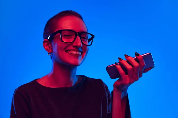 Jeune fille émotionnelle avec coupe buzzcut coiffure en utilisant le téléphone isolé sur fond bleu foncé dans la lumière au néon. Concept d'émotions — Photo