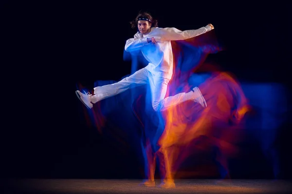 Flexibilní muž v bílém sportovním obleku tančící hip-hop izolovaný na tmavém pozadí ve smíšeném neonovém světle. Mládež kultura, hip-hop, pohyb, styl a móda, akce. — Stock fotografie