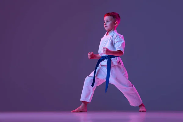 Dinamikus portré a kölyökről, fiatal férfi taekwondo, karate sportolók dobokokban, akik lila háttérrel elszigetelt alapmozdulatokat csinálnak a neonban. A sport, a harcművészetek fogalma — Stock Fotó
