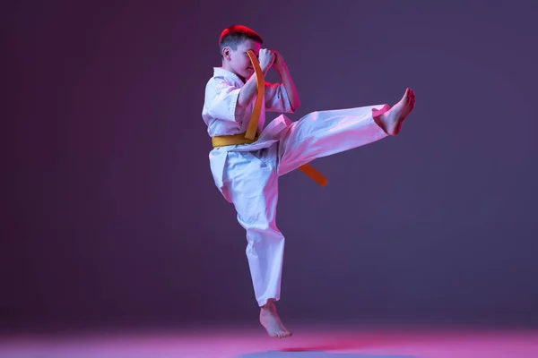 Retrato de niño deportivo, taekwondo masculino, atletas de karate en doboks haciendo movimientos básicos aislados sobre fondo púrpura en neón. Concepto de deporte, artes marciales —  Fotos de Stock
