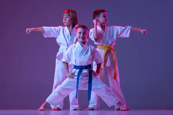Tres niños deportivos, niños pequeños, taekwondo o atletas de karate en doboks posando aislados sobre un fondo de color muy peri en neón. Concepto de deporte, artes marciales — Foto de Stock