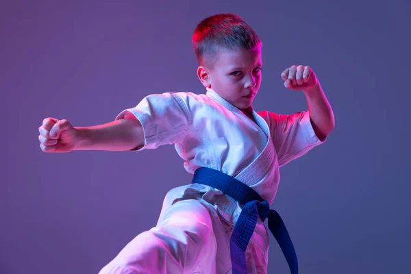 Retrato dinámico de niño, joven taekwondo masculino, atletas de karate en doboks haciendo movimientos básicos aislados sobre fondo púrpura en neón. Concepto de deporte, artes marciales — Foto de Stock