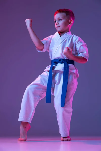 Retrato de niño deportivo, taekwondo masculino, atletas de karate en doboks haciendo movimientos básicos aislados sobre fondo púrpura en neón. Concepto de deporte, artes marciales — Foto de Stock