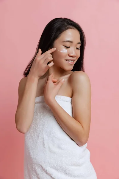 Retrato de jovem mulher bonita em toalha de banho branca rosto hidratante com creme isolado sobre fundo de estúdio rosa — Fotografia de Stock