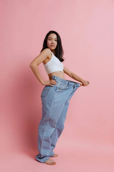 Retrato completo de bela mulher magra posando em jeans enormes isolados sobre fundo de estúdio rosa. Conceito de perda de peso — Fotografia de Stock