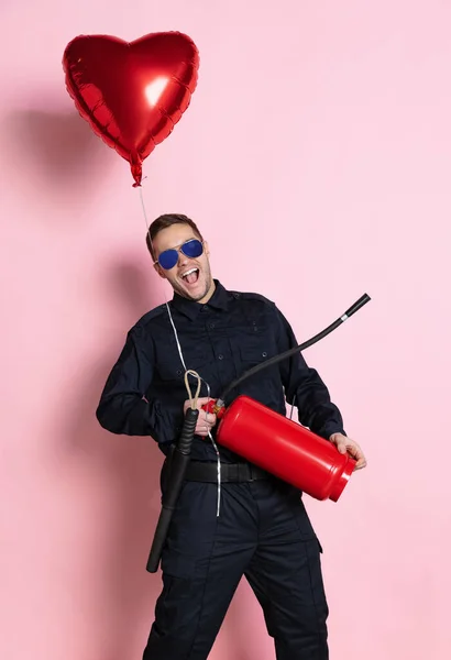 Κωμικό πορτρέτο του νεαρού γοητευτικού αστυνομικού που διασκεδάζει με μπαλόνι σε σχήμα καρδιάς και πυροσβεστήρα που απομονώνονται σε ροζ φόντο. Άγιος Βαλεντίνος ημέρα — Φωτογραφία Αρχείου