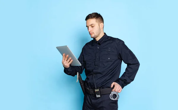 青い背景に隔離されたタブレットを使用して黒の制服を着た若い男、警官の肖像画。仕事、ケア、法律、秩序の概念. — ストック写真