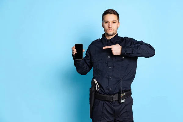 Portret młodego człowieka, policjant w czarnym mundurze z telefonem odizolowanym na niebieskim tle. Pojęcie pracy, kariery, prawa i porządku. — Zdjęcie stockowe