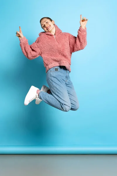 年轻女孩的动态肖像，穿着粉色帽衫的短发学生在蓝色背景下跳跃。情感、音乐的概念 — 图库照片