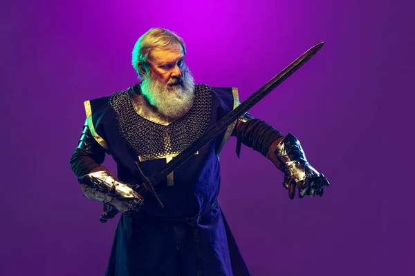 Retrato do homem grisalho sênior, bravo e brutal guerreiro medieval ou cavaleiro de armadura com espada isolada sobre fundo púrpura em néon. Comparação de eras — Fotografia de Stock