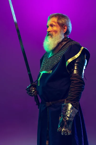 Retrato do homem grisalho sênior, bravo e brutal guerreiro medieval ou cavaleiro de armadura com espada isolada sobre fundo púrpura em néon. Comparação de eras — Fotografia de Stock