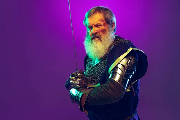 Porträt eines älteren graubärtigen Mannes, tapferer und brutaler mittelalterlicher Krieger oder Ritter in Rüstung mit Schwert isoliert über violettem Hintergrund in Neon. Epochenvergleich — Stockfoto
