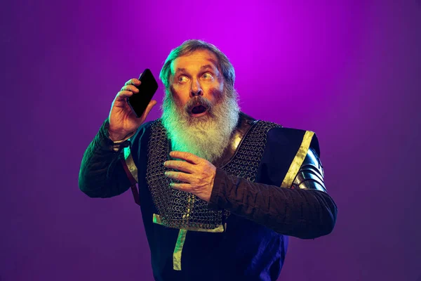 Retrato cómico del hombre mayor de barba gris en imagen de guerrero medieval en armadura usando smartphone aislado sobre fondo púrpura en neón. Comparación de épocas — Foto de Stock