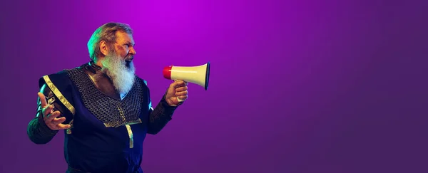 O Flyer. Engraçado velho homem de barba cinza em imagem de guerreiro medieval ou cavaleiro em traje período gritando em megafone isolado o fundo roxo em néon. Comparação de eras — Fotografia de Stock