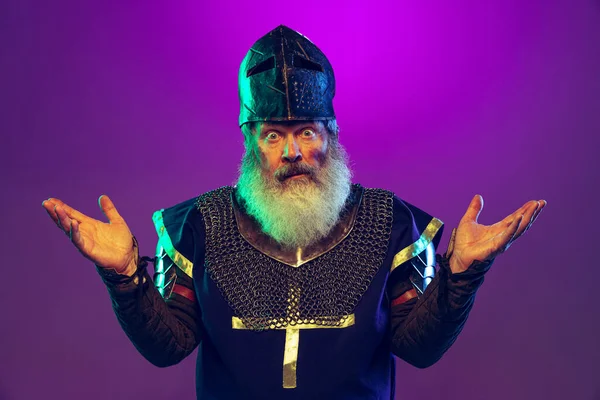 Surprised senior grey-bearded man in image of medieval warrior or knight in armor with sword isolated over purple background in neon. Comparison of eras — Stock Photo, Image