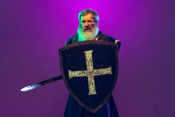 Komiczny portret starszego siwobrodego mężczyzny na obraz średniowiecznego wojownika lub rycerza w zbroi z mieczem odizolowanym na fioletowym tle w neonie. Porównanie epok — Zdjęcie stockowe
