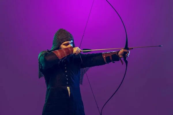 Студійний знімок молодого бородатого чоловіка на зображенні середньовічного воїна, стрільба з лука в ланцюговій броні ізольовані на фіолетовому фоні в неоні. Порівняння епох — стокове фото