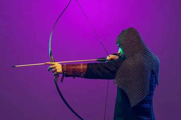 Estúdio tiro de jovem barbudo na imagem de guerreiro medieval, tiro com arco em cadeia armadura isolada sobre fundo roxo em néon. Comparação de eras — Fotografia de Stock