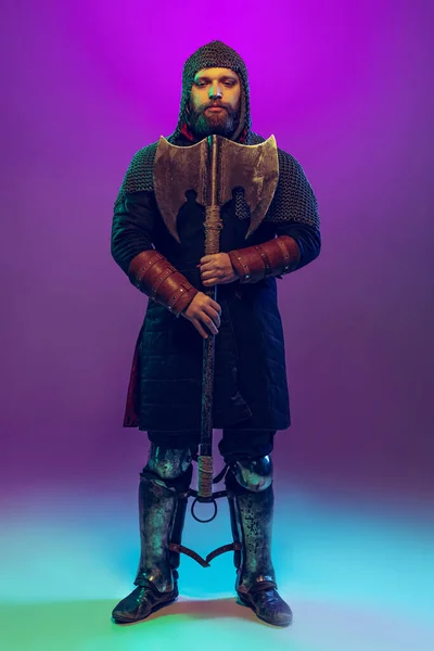 Studioaufnahme eines jungen bärtigen Mannes im Bild eines mittelalterlichen Kriegers, Bogenschießen in Kettenrüstung isoliert über violettem Hintergrund in Neon. Epochenvergleich — Stockfoto