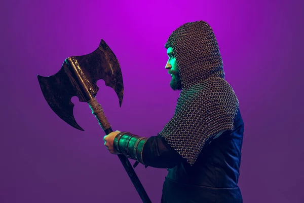 Studioaufnahme eines jungen bärtigen Mannes im Bild eines mittelalterlichen Kriegers, Bogenschießen in Kettenrüstung mit Axt isoliert über violettem Hintergrund in Neon. Epochenvergleich — Stockfoto