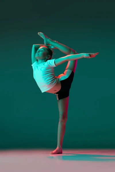 Piccola ragazza flessibile, ginnastica ritmica formazione artista isolato su sfondo studio verde in luce rosa neon. Grace in movimento, azione. Esercizi di flessibilità. — Foto Stock