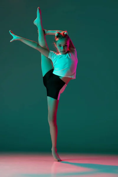 Pequena menina flexível, ginástica rítmica formação artista isolado no fundo do estúdio verde em luz rosa néon. Graça em movimento, acção. Fazendo exercícios em flexibilidade. — Fotografia de Stock
