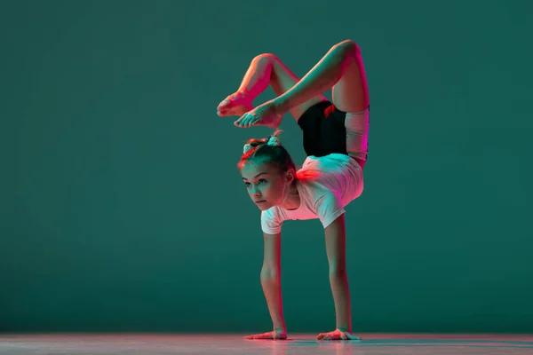 Handstand. Little flexible girl, rhythmic gymnastics artist training isolated on green studio background in neon pink light. Grace in motion, action. Doing exercises in flexibility. — Stock Photo, Image