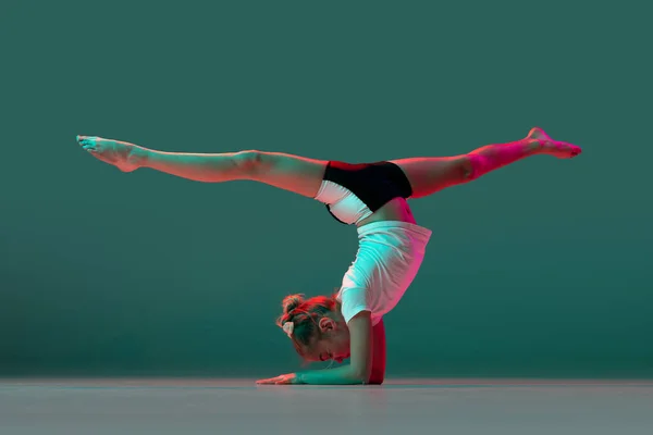 Handstöd. Liten flexibel flicka, rytmisk gymnastik artist utbildning isolerad på grön studio bakgrund i neon rosa ljus. Nåd i rörelse, handling. Göra övningar i flexibilitet. — Stockfoto
