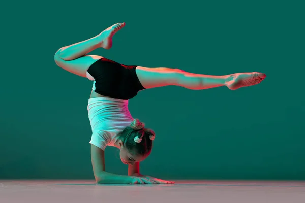 Handstand. Little flexible girl, rhythmic gymnastics artist training isolated on green studio background in neon pink light. Grace in motion, action. Doing exercises in flexibility. — Stock Photo, Image