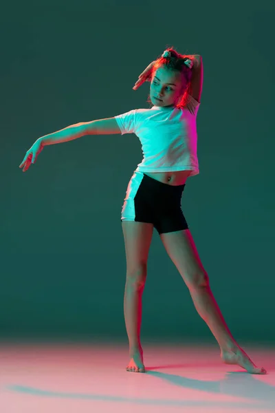 Kleines flexibles Mädchen, rhythmische Turnerin, die isoliert auf grünem Studiohintergrund in neonrosa Licht trainiert. Gnade in Bewegung, Aktion. Übungen in der Beweglichkeit. — Stockfoto