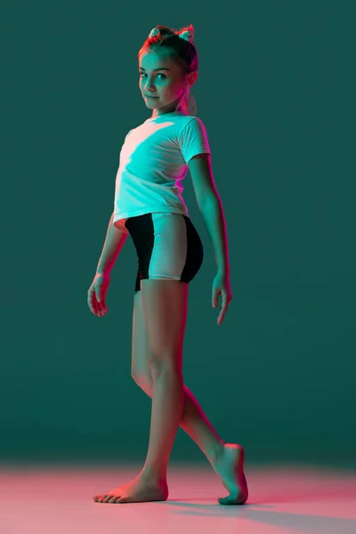 Portret pięknej dziewczynki, artystki gimnastyki rytmicznej w mundurze sportowym, pozującej na zielonym tle studia w neonowym, różowym świetle. Sport, dzieciństwo, koncepcja studiów — Zdjęcie stockowe