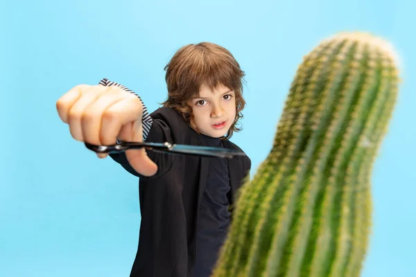 Cortando cactus. Retrato cómico de niño pequeño, niño con una enorme chaqueta negra como peluquero divirtiéndose aislado sobre fondo azul. — Foto de Stock